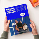 Blaues Magazin „Harvard Business manager“ mit weißem Titel „Das Wissen der Besten“, gehalten von zwei Händen in grauen Strickärmeln auf weißer Fläche; oben rechts liegt ein orangefarbenes Tablett. Sichtbare Texte: „Produkte: Wie Game Boy und Crocs zu Ikonen wurden“, „Künstliche Intelligenz: Warum Ikea Chatbots umgarnt“, „Marketing: Zahlen sich immersive Erlebnisse aus?“, „Selbstmanagement: Wann es Zeit ist, Prioritäten neu zu setzen“, „Jetzt gib schon auf…“, „Nö.“ und „Verhandeln am Limit“.