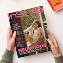 Schräg auf einer weißen Fläche gehaltene Modezeitschrift „InStyle“ mit schwarzem Cover und pinker Typografie; darauf eine weibliche Person mit braunem Haar in hellem Oberteil vor einem Fenster. Lesbare Aufschriften sind „47 TREND-SHOES“, „NEUE MODE“, „SKIN-TONE HAIR“ und „LESLIE MANN“; zwei Hände mit grauen Strickärmeln halten das Magazin, oben rechts liegt ein orangefarbenes rechteckiges Objekt.