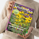 Person ohne sichtbares Gesicht hält mit beiden Händen ein Magazin mit violettem Rand und glänzendem Cover vor einem beigefarbenen Strickpullover. Der Titel lautet „Kraut&Rüben – Schöner. Wilder. Biogarten.“; sichtbar sind gelbe Blüten vor violett-grünem Hintergrund sowie die Texte „Cool-Flower-Methode: Jetzt säen, im Frühling strahlen“, „Beste Aussichten“ und „Gärtnern im Schein der späten Sonne“.