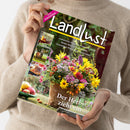 Weibliche Person im beigen Strickpullover hält eine schräg fotografierte Zeitschrift mit dem Titel „Landlust“ und dem Hinweis „NEUE STRICKIDEEN“. Das Cover zeigt einen bunten Blumenstrauß im Topf, Äpfel, einen Hirsch und weitere Naturmotive; lesbare Texte sind „Laubsammler: Aufsatz für Schubkarren bauen“, „Basteln: Blüten aus Klopapier“ und „Der Herbst zieht ein“.