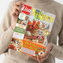 Person in beigem Strickpullover hält eine schräg fotografierte Zeitschrift mit dem Titel „Lisa Wohnen & Dekorieren“. Das Cover zeigt orange Herbstblumen im Weidenkorb, Kürbisse, herbstliche Dekoration und Wohnaccessoires; lesbare Texte sind „58 kreative Herbst-Ideen“, „Dekolust im Herbst“, „Wohlfühl-Mix“ und „30 clevere Tipps für einen einladenden Flur“.