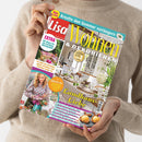 Weibliche Person im beigefarbenen Strickpullover hält eine schräg ausgerichtete Wohnzeitschrift mit türkisfarbenem, rotem und violettem Cover. Sichtbare Texte: „Kreativ den Sommer verlängern“, „Wohnen & Dekorieren“, „Landhaus-Liebe“ und „EXTRA 60 Ideen für ein leichtes Wohngefühl“; abgebildet sind ein dekorierter Esstisch, Blumen sowie eine Frau mit Blumenstrauß.