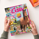 Schräg gehaltenes, farbiges Pferdemagazin „Lissy“ mit großem braunem Pferdefoto auf hellblauem Hintergrund, pinkem Rand und zwei Händen an den Seiten. Sichtbare Texte: „COOLER GEWINN!“, „FOTOROMAN“, „REPORT“, „BACKSTAGE BEIM Reitturnier“, „DIY Pferdestärke ABKÜHLUNG“, „POSTER ZUM VERLIEBEN“, „FUN ZUM WIEHERN KOMISCH“, „TEST Welcher Pferde-Typ passt zu dir?“ und „Retro Comic“; oben rechts liegt ein orangefarbenes Schreibbrett.