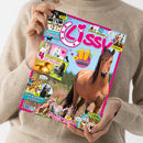 Weibliche Person im beigen Strickpullover hält eine schräg fotografierte, farbenfrohe „Lissi“-Pferdezeitschrift mit pinkem Rand. Das Cover zeigt mehrere Pferdefotos, eine große Pferdeaufnahme, Comicbilder und Texte wie „REPORT“, „BACKSTAGE BEIM Reitturnier“, „COOLER GEWINN!“, „FOTOROMAN“, „4 POSTER ZUM VERLIEBEN“, „DIY Pferdestärke ABKÜHLUNG“, „ZUM WIEHERN KOMISCH“ und „Retro Comic“.