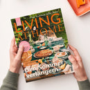 Aufgeschlagenes Magazin „LIVING AT HOME“ mit dem Titel „Mein Zuhause voller Ideen“ und dem Hauptthema „Der Sommer verlängern“. Das Cover zeigt einen gedeckten Gartentisch mit Kuchen, Getränken und Blumen; sichtbar sind außerdem die Texte „Citytrip Porto“, „Spätsommer-Glück: Dahlien, Zinnien und Sonnenblumen“ sowie zwei Hände, die das Magazin halten.