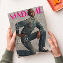Schräg gehaltene graue Modezeitschrift „MADAME“ mit männlicher Person in blauem Jeanshemd, Jeans und schwarzen Stiefeln auf dem Cover; zwei Hände halten das Magazin. Sichtbare Texte: „ENDLICH FIT“, „Leicht & straff mit dem richtigen Krafttraining“, „WELT·RÄUME“, „Die jungen Meister des Interior-Designs“, „RELAX!“ und „So lässig ist die neue Mode“; oben rechts liegt ein orangefarbenes Notizbuch.