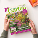 Schräg gehaltenes Gartenmagazin „mein schöner Garten“ mit grünem Titel, Blumenbeet auf dem Cover und zwei Händen an den unteren Seitenkanten; oben rechts liegt ein orangefarbenes Gartengerät. Sichtbare Texte: „Europas größtes Gartenmagazin“, „Klimaschutz + Artenvielfalt“, „Kein Garten ohne Hecke“, „Zucchini & Kürbis – Beet auf den Tisch“ und „Blütenglück mit pflegeleichten Fetthennen“.