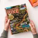 Schräg auf einem weißen Tisch liegendes, von zwei Händen gehaltenes mountainBIKE-Magazin mit glänzendem Cover. Sichtbare Texte: „mountainBIKE“, „E-MTB“, „Kupplungsträger“, „Dreier-Träger im Test“, „28 Seiten Reise“, „14 XC-Reifen im Labor- & Praxistest“, „BOCK auf SPEED“, „9 QWM-Bikes im Test“ und „E-CVT“. Das Cover zeigt einen bewaldeten Mountainbike-Trail mit fahrender Person und Fahrrädern auf einem Autoträger.