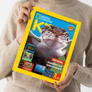 Weibliche Person im hellbeigen Strickpullover hält ein schräg ausgerichtetes Magazin mit gelbem Rand. Das Cover von „NATIONAL GEOGRAPHIC“ zeigt einen braun-weiß gefleckten Gecko; lesbare Texte sind „COOLE FAKTEN“, „Verrückt, aber wahr!“, „GECKOS: Kopfüber auf Insektenjagd!“, „2 tolle POSTER!“ sowie „Schiffswracks“, „Sommer“ und „Schokolade“.