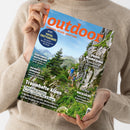 Eine weibliche Person in beigefarbenem Strickpullover hält schräg eine Zeitschrift mit glänzendem Cover. Darauf stehen „Outdoor“, „Wandern. Reisen. Abenteuer.“, „Total trittsicher! Die besten Wanderschuhe für die Berge“, „Traumhafte Alpen“, „Japans Wanderjuwel“ und „Im Check: Rucksäcke“; abgebildet sind zwei wandernde Personen in einer grünen, felsigen Berglandschaft.