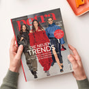 Aufgeschlagenes Magazin „petra“, von zwei Händen über einer weißen Oberfläche gehalten. Das Cover zeigt drei weibliche Models in schwarzer, roter und blauer Kleidung sowie die Texte „Mein Leben! Mein Luxus!“, „DIE NEUEN TRENDS“, „PREMIUM-HEALTH: DER NEUE STOFFWECHSEL-CODE“, „STYLISCHE FRISUREN“, „BRETONISCHE GEHEIMNISSE“ und „VON MAILAND BIS PARIS“.