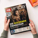 Schräg gehaltenes Magazin „PM HISTORY“ mit schwarzem Cover, zwei Händen und grauen Strickärmeln. Das Titelbild zeigt eine Frau mit dunklem Haar, Stirnband und Perlenkette in historischer Kleidung; lesbare Texte: „Europas größtes Monatsmagazin für Geschichte“, „DIE GOLDENEN ZWANZIGER“, „Was fasziniert uns heute noch an diesem Jahrzehnt zwischen Rausch und Ruin?“, „Mehr als nur Muster“ und „BUCHKUNST“.