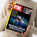 Weibliche Person im cremefarbenen Strickpullover hält ein schräg fotografiertes Magazin mit schwarzem Cover und gelbem Rand. Sichtbare Texte: „PM. Das Wissensmagazin“, „Macht KI dumm?“, „Wein-Archäologie“, „Löschroboter“, „Hightech-Klos analysieren unsere Gesundheit“ sowie „Lithium, Kobalt, Wolfram – Bodenschätze Made in Germany“.