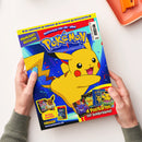 Zwei menschliche Hände in grauen Strickärmeln halten eine rechteckige Pokémon-Zeitschrift vor weißem Hintergrund. Das farbenfrohe Cover zeigt Pikachu auf blauem Grund sowie die Texte „3D-Anhänger“, „FUNPACK“, „XL-POSTER“, „POSTKARTEN“, „HIGHLIGHT-AUSGABE“, „Schnapp sie dir alle!“, „4 Postkarten mit Goldprägung!“, „Mit Mega-Gewinnspiel!“, „Panini“ und „www.pokemon.de“.