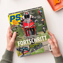 Aufgeschlagenes Magazin „PS Sport motorrad magazin“, schräg von oben von zwei Händen gehalten. Das Cover zeigt eine Person mit Helm auf einem roten Sportmotorrad, mehrere weitere Motorräder sowie die Texte „ZONKOS ER-FAHRUNGEN: WIE ICH MOTORRADJOURNALIST WURDE“, „SPORTSCHRIFT“ und „Feine Zweitakt-Renner“; oben rechts liegt ein orangefarbenes Objekt.
