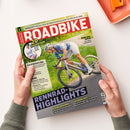 Auf weißem Untergrund halten zwei Hände ein schräg liegendes Magazin „ROADBIKE“ mit dem Titel „RENNRAD-HIGHLIGHTS“. Das Cover zeigt eine Radfahrerin mit weißem Helm, blauem Trikot und weißer Radhose auf einem violetten Rennrad; sichtbar sind außerdem „20 RADHOSEN“, „Werkstatt: Was tun, wenn die Elektroschaltung streikt? So hilfst du dir selbst“ und „Räder des Jahres“. Oben rechts liegt ein orangefarbenes Utensil.