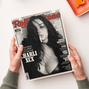 Schräg auf weißer Fläche gehaltenes Magazin „Rolling Stone“ mit schwarz-weißem Porträt einer Frau mit langen dunklen Haaren und Zigarette. Sichtbare Texte: „Popkultur & mehr“, „August 2026“, „Die 50 besten Pop-Alben“ sowie „Madonna“, „Daft Punk“, „Beyoncé“, „Billie Eilish“, „Bad Bunny“, „Phoebe Bridgers“, „Sting“, „James Ford“ und „Kanye West“; zwei Hände und ein orangefarbenes Objekt sind sichtbar.