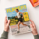 Schräg liegendes Magazin „RUNNERS WORLD“ mit blauem Cover, gelber und roter Schrift sowie Titelbild einer weiblichen Läuferin in weißem Oberteil und schwarzer Shorts auf steinigem Untergrund. Sichtbare Texte: „DAS GRÖSSTE LAUFMAGAZIN DER WELT“, „Diese Salate machen wirklich schneller“, „Stabiler laufen“, „Home-Workout für eine starke Hüfte“ und „32 SEITEN TRAIL-SPECIAL“; zwei Hände halten das Magazin.