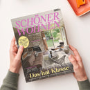 Wohnmagazin „SCHÖNER WOHNEN“ liegt schräg auf einer weißen Fläche und wird von zwei Händen mit grün-grauen Strickärmeln gehalten. Das Cover zeigt ein Wohnzimmer mit Steinkamin, braunem Sofa, schwarzem Lounge-Sessel, rundem Tisch und Blick in einen Garten; sichtbare Texte: „EUROPAS GRÖSSTES WOHNMAGAZIN“, „FACELIFT FÜR DIE KÜCHE“, „RAUMWUNDER“ und „Das halbe Klasse“.