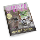 Schräg fotografiertes Cover der Wohnzeitschrift „SCHÖNER WOHNEN“ mit pinkem Titel und dem Zusatz „EUROPAS GRÖSSTES WOHNMAGAZIN“. Abgebildet ist ein modernes Wohnzimmer mit Steinkamin, Sofa, Ledersessel, Couchtisch, Teppich und großen Fenstern; sichtbare Texte: „FACELIFT FÜR DIE KÜCHE“, „RAUMWUNDER“, „BETON, HOLZ, FARBE“ und „Das halb Klasse“.