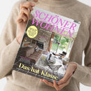 Person in beigefarbenem Strickpullover hält ein schräg fotografiertes Magazincover mit dem pinken Titel „SCHÖNER WOHNEN“ und dem Text „EUROPAS GRÖSSTES WOHNMAGAZIN“. Sichtbar sind außerdem „FACELIFT FÜR DIE KÜCHE“, „RAUMWUNDER“, „ÜBERRASCHUNG!“ und „Das hat Klasse“; das Cover zeigt ein Wohnzimmer mit Kamin, Sofa, Sesseln und großen Fenstern.