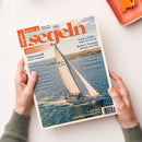 Auf weißer Fläche hält eine Person mit beiden Händen ein leicht schräg liegendes Magazin mit orangefarbenem Titel „segeln“ und dem Text „Fahrten- und Freizeitsegeln“, „Juli August 2026“, „Nach Gefühl … Segeln ohne Technik“ sowie „Recht & Technik“. Das Cover zeigt ein Segelboot auf blauem Meer, weitere Texte wie „Dänemark“, „Decksalon“ und „Gebrauchtboote“; oben rechts liegt ein orangefarbenes rechteckiges Objekt.