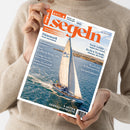 Eine Person ohne sichtbares Gesicht hält schräg eine Zeitschrift mit dem Titel „Segeln“ und orange-weißem Cover. Das Titelbild zeigt eine weiße Segelyacht auf blauem Meer; lesbare Texte sind „100% Fahrten- und Freizeitseglern“, „Dänemark – Fünen und Seeland“, „Deckssalon im Eigenbau“ und „8 Seiten Gebrauchtboote“.