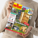 Person in beigem Strickpullover hält eine schräg fotografierte Zeitschrift mit gelbem Titel „selbst ist der Mann“ und dem Untertitel „Das Do-it-yourself-Magazin“. Das Cover zeigt Gartenhäuser, eine Person mit Laubsauger, einen Mann beim Bauen, Rostschutzarbeiten und eine Kreissäge; sichtbare Texte sind „Gartenhäuser – 2 Projekte selber bauen“, „Laub-Saugbläser im Praxistest“, „Anti-Rost: Geländer aus Metall streichen“ und „Großes Saunahaus“.