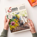 Eine Person hält das Magazin „Servus“ mit dem Untertitel „einfach gut leben“ vor hellem Hintergrund. Das Cover zeigt eine grüne Gießkanne, gerollte weiße Tücher und bunte Blumen auf einer Holzoberfläche; sichtbare Texte sind „BALSAM FÜR DIE SEELE“, „Wie uns die Natur mehr Lebensfreude schenkt“, „Der beste Kaiserschmarrn der Welt“ sowie „Paul Pizzera und das Zniachtl“.