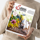 Eine Person im beigen Strickpullover hält eine schräg ausgerichtete weiße Zeitschrift mit dem Titel „Genus“ und dem Untertitel „einfach gut leben“. Das Cover zeigt bunte Blumen, eine gelbe Kanne und gerollte Handtücher; lesbare Texte: „Balsam für die Seele“, „Der beste Käsemann der Welt“ und „Kulinarische Spurensuche hoch über Bad Gastein“.