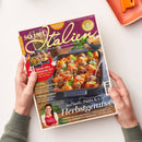 Schräg gehaltenes Kochmagazin „so isst Italien“ mit violettem Titelbild, zwei Händen und grauen Ärmeln vor weißem Hintergrund. Sichtbare Texte: „82 geniale Rezepte“, „Für Ihre italienischen Momente“, „Rigatoni al forno mit Hackbällchen“, „41 Saison-Hits“, „Kürbis: 3-mal köstlich“, „Ofengerichte“ und „Pasta & Co.“; abgebildet sind ein Auflauf mit Pasta, Hackbällchen und Basilikum sowie ein orangefarbenes Küchengerät.