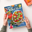 Schräg von oben fotografierte Zeitschrift „so ist Italien“, von zwei Händen über einer hellen Fläche gehalten; daneben liegt oben rechts ein orangefarbenes Tablett. Das Cover zeigt einen bunten Teller mit Gemüse und gefüllten Rouladen sowie eine kleine Frauendarstellung und trägt unter anderem die Texte „94 geniale Rezepte“, „Italienische Urlaubsküche“, „Pomodori“ und „Dolce Vita auf dem Teller!“.