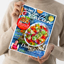 Person im beigen Strickpullover hält eine diagonal ausgerichtete, glänzende Kochzeitschrift mit blau strukturierter Titelseite. Sichtbare Texte: „So is(s)t Italien“, „94 geniale Rezepte“, „30 x schnell und einfach kochen“, „Italienische Urlaubsküche“, „Pomodori!“ und „Dolce vita auf dem Teller!“; abgebildet sind ein bunter Tomaten-Mozzarella-Salat, weitere Speisen sowie ein Porträt einer Frau.