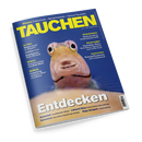 Schräg fotografiertes, blau-gelbes Tauchmagazin „TAUCHEN“ mit glänzendem Umschlag und weißem Seitenrand auf schwarzem Hintergrund. Zentral ist ein gelblich-brauner Fisch mit großen schwarzen Augen und geöffnetem Maul abgebildet; sichtbare Texte sind „Wissen: Fotoschule – Sprechstunde – Apnoe-Tauchen“, „Geiseltalsee: Spektakulärer Unterwasserwald“, „Atemregler Techniker-Kurs“ und „Entdecken“.