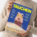 Eine Person ohne sichtbares Gesicht hält ein schräg ausgerichtetes, glänzendes Magazin mit blauem Cover und großem gelbem Titel „TAUCHEN“ vor einem beigefarbenen Strickpullover. Abgebildet ist ein großformatiger Fischkopf; sichtbare Texte nennen „Wissen: Fotoschule – Sprechstunde – Apnoe-Tauchen“, „Entdecken“, „Sulawesi“, „Lahami Bay“, „Bahamas“ und „Raja Ampat“.