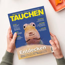 Auf weißem Untergrund hält eine Person mit beiden Händen ein schräg liegendes Magazin mit blauem Cover und großem gelbem Titel „TAUCHEN“. Abgebildet ist ein nahes Unterwasserfoto eines Fisches; lesbare Texte sind „Wissen: Fotoschule – Sprechstunde – Apnoe-Tauchen“, „Entdecken“ und „Geiseltalsee Spektakulärer Unterwasserwald“. Oben rechts liegt teilweise ein orangefarbenes rechteckiges Objekt.