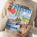 Eine Person ohne sichtbares Gesicht hält ein schräg fotografiertes Magazin mit braunem Umschlag und blauem Titel „Zuhause Wohnen“ vor einem beigefarbenen Strickpullover. Das Cover zeigt eine helle Wohnterrasse am Meer mit weißen Polstermöbeln und Pflanzen sowie die Texte „Wie wohnen glücklich macht“, „Genuss weltweit“, „12 Regale“, „Blickfang Bilder“, „Leben wie im Urlaub“ und „Kaffee & Eis“.